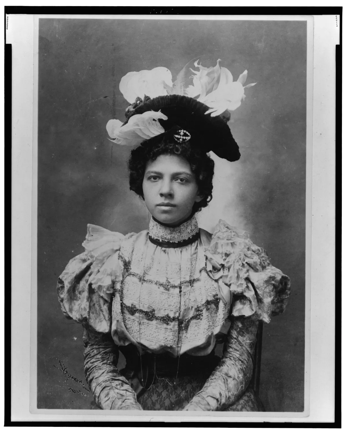 A portrait of an African American woman wearing an ornate hat adorned with feathers, lace, and a brooch, dressed in a detailed blouse with puffed sleeves.