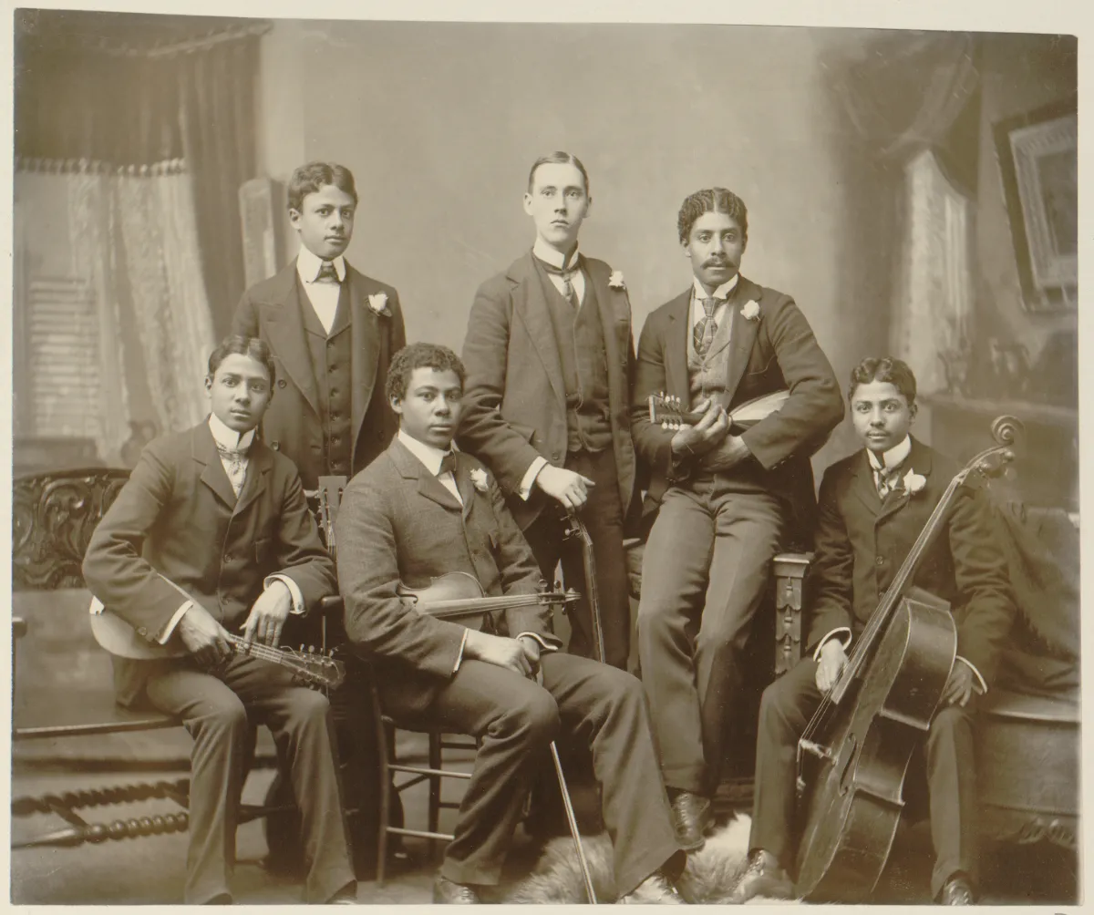 A group portrait featuring five African American men, Thomas E. Askew’s sons, and a neighbor, posing with their musical instruments, including violins, a cello, and a mandolin, dressed in formal suits.