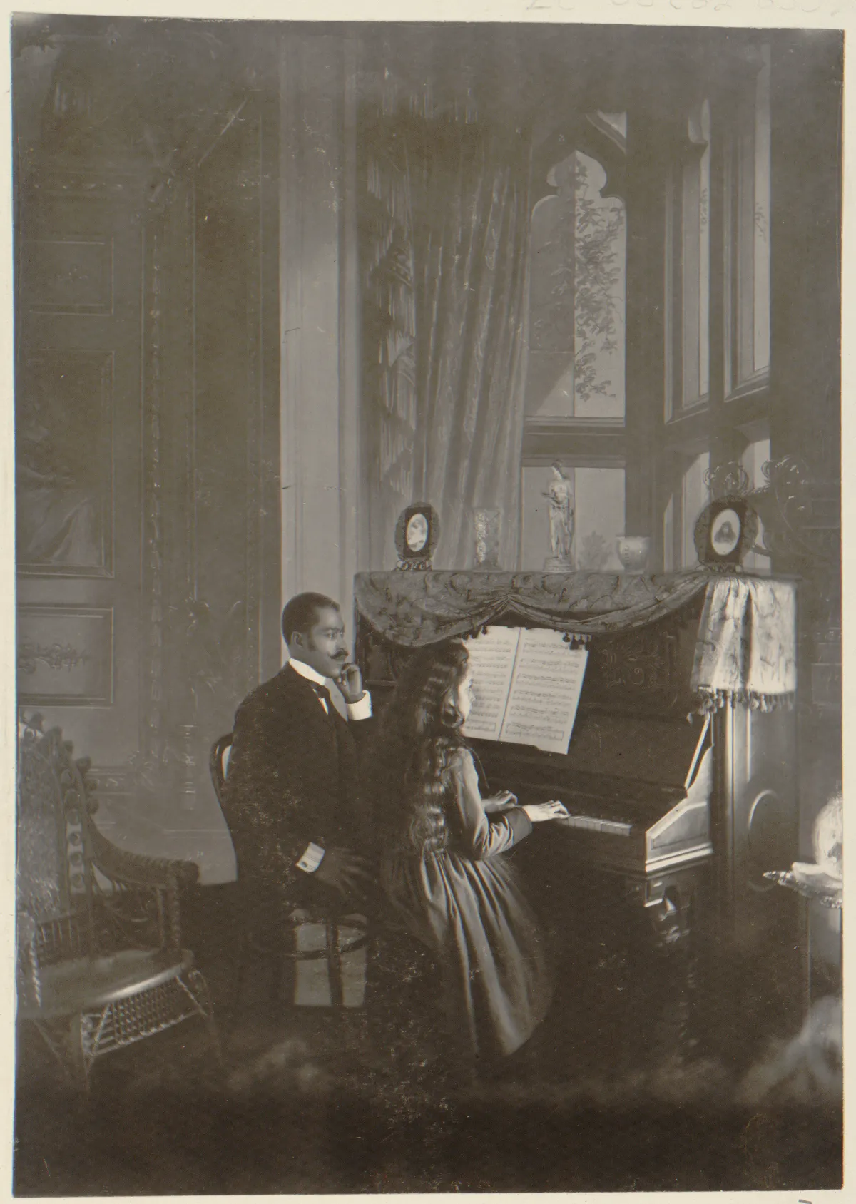 An African American man giving a piano lesson to a young woman, seated in a dimly lit parlor with decorative furnishings, including a piano draped with fabric.