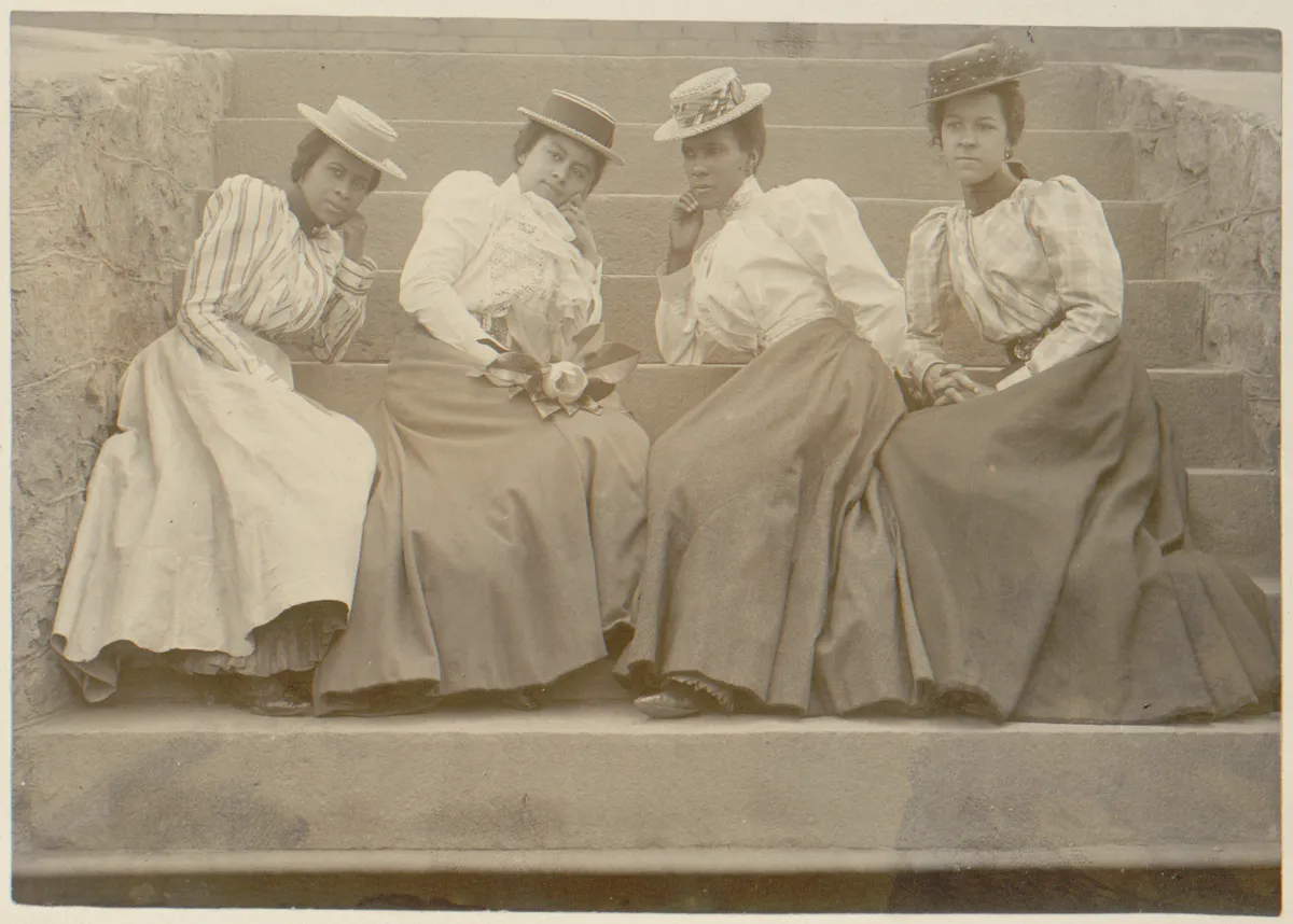 Four African American women dressed in long skirts and blouses, wearing hats, sitting on the steps of a building at Atlanta University.