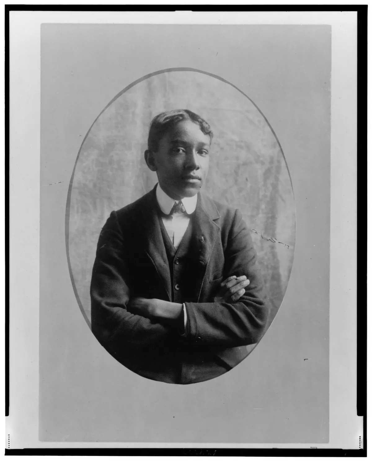 A young African American man posing with his arms crossed, dressed in a dark suit and tie, in an oval-shaped portrait frame.