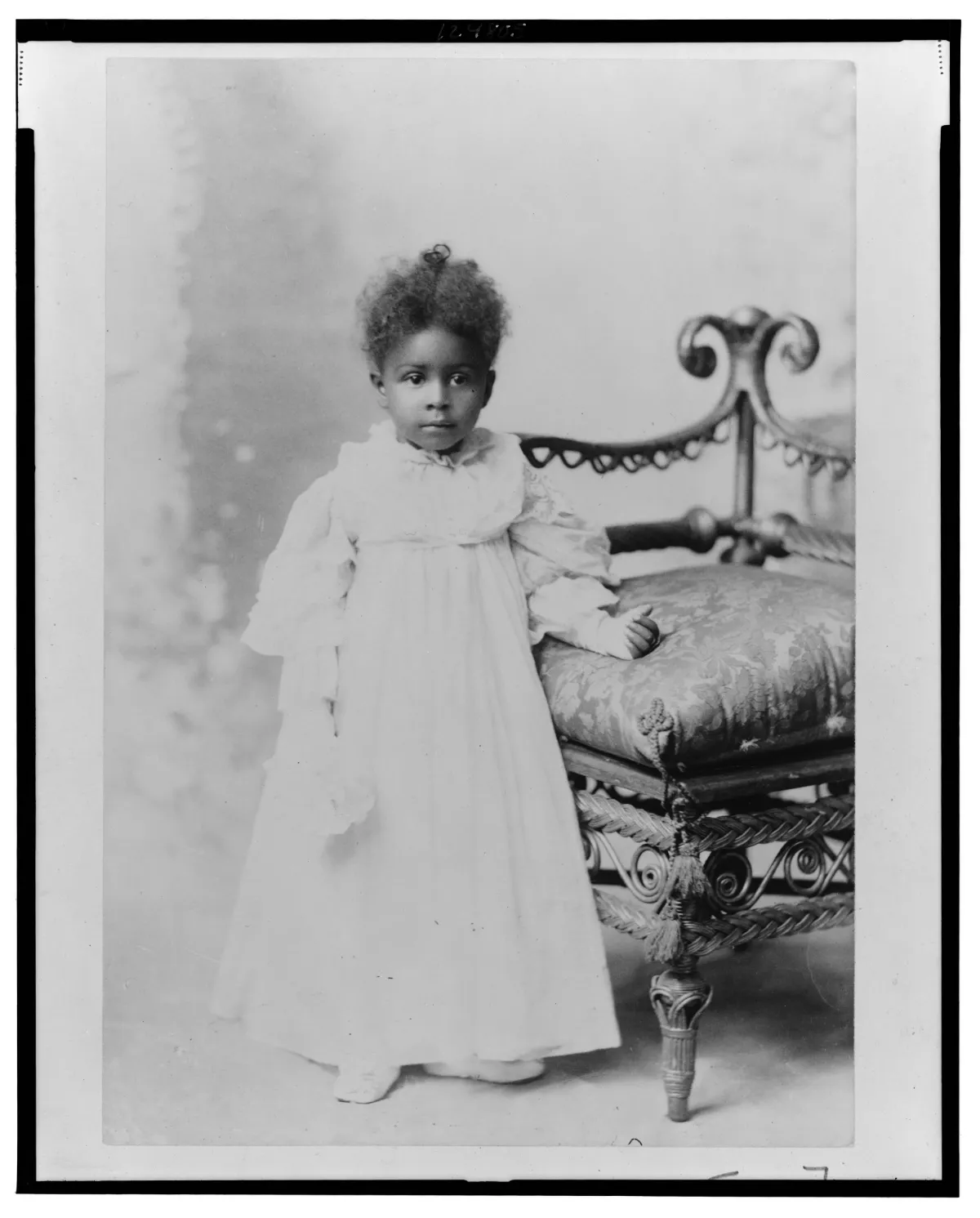 A portrait of a young African American girl wearing a long white dress, standing beside an intricately carved wooden chair, with a neutral studio backdrop.