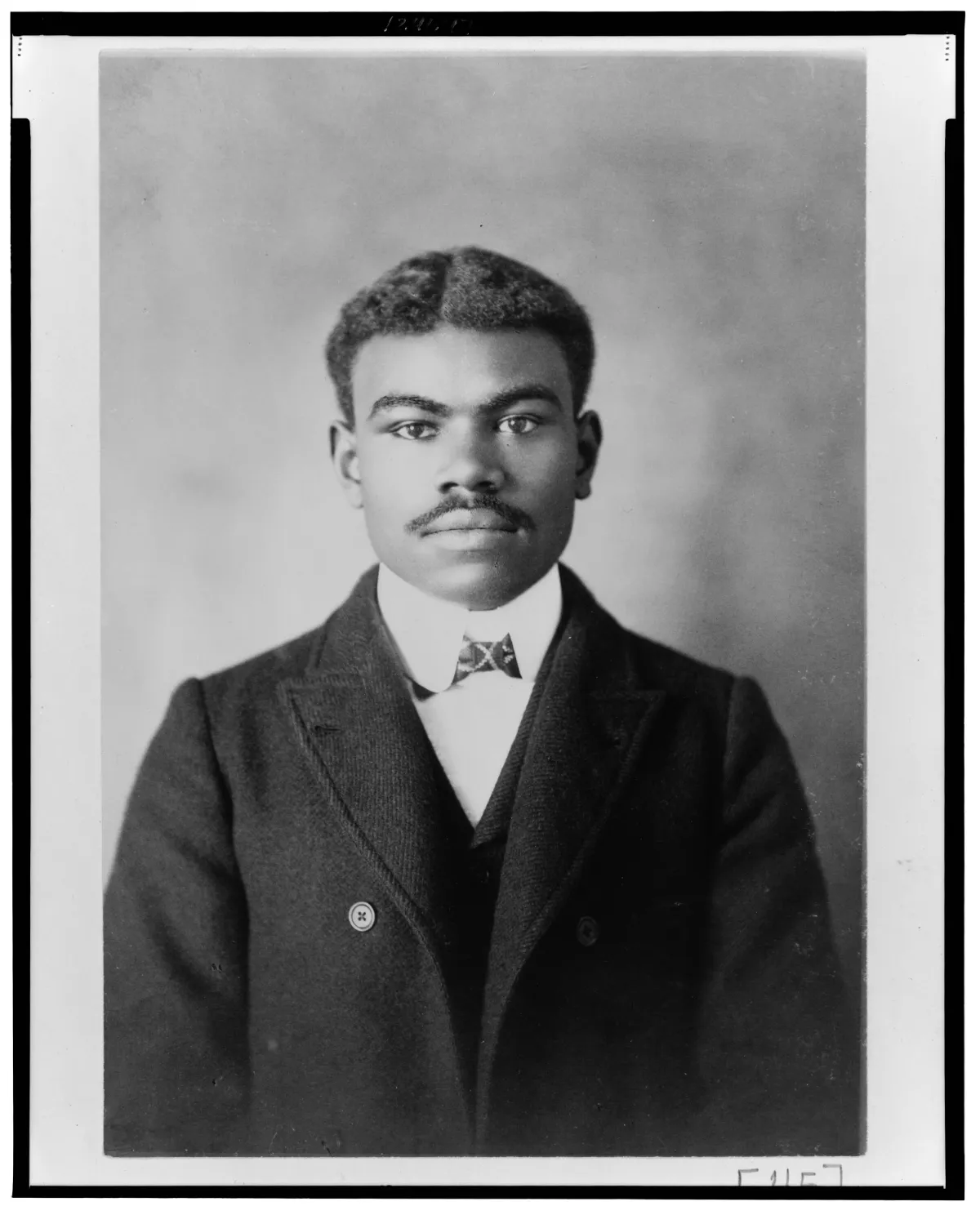 A formal portrait of an African American man dressed in a dark double-breasted coat, white shirt, and patterned bow tie, with a neutral studio backdrop.