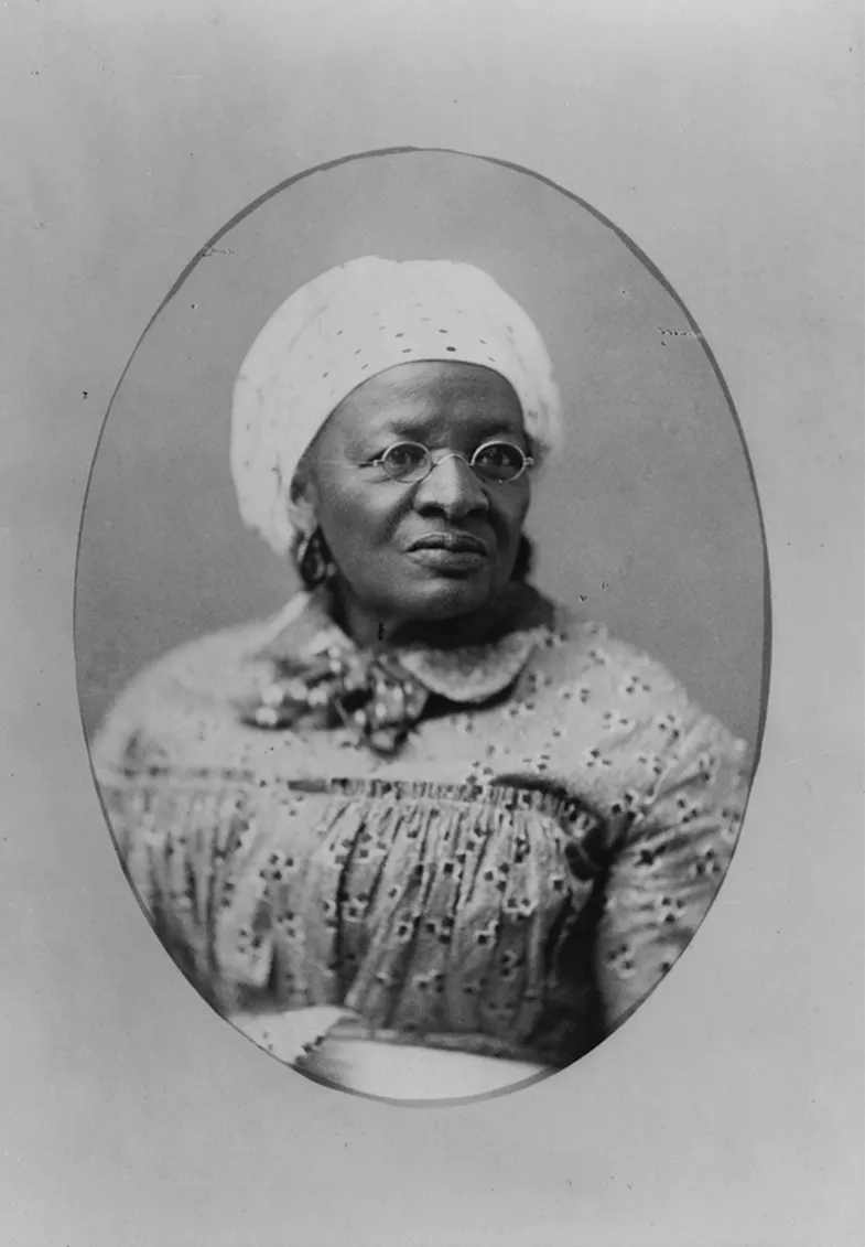 Portrait of an elderly African American woman wearing glasses, a patterned dress with a high neck, and a cap, conveying a sense of dignity and resilience.