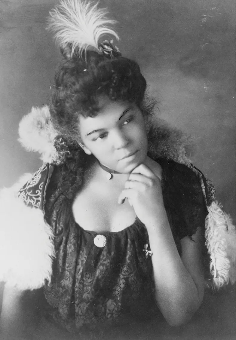 Portrait of a young African American woman with a feathered headpiece, dressed in a decorative lace top, her hand under her chin as she gazes thoughtfully to the side.