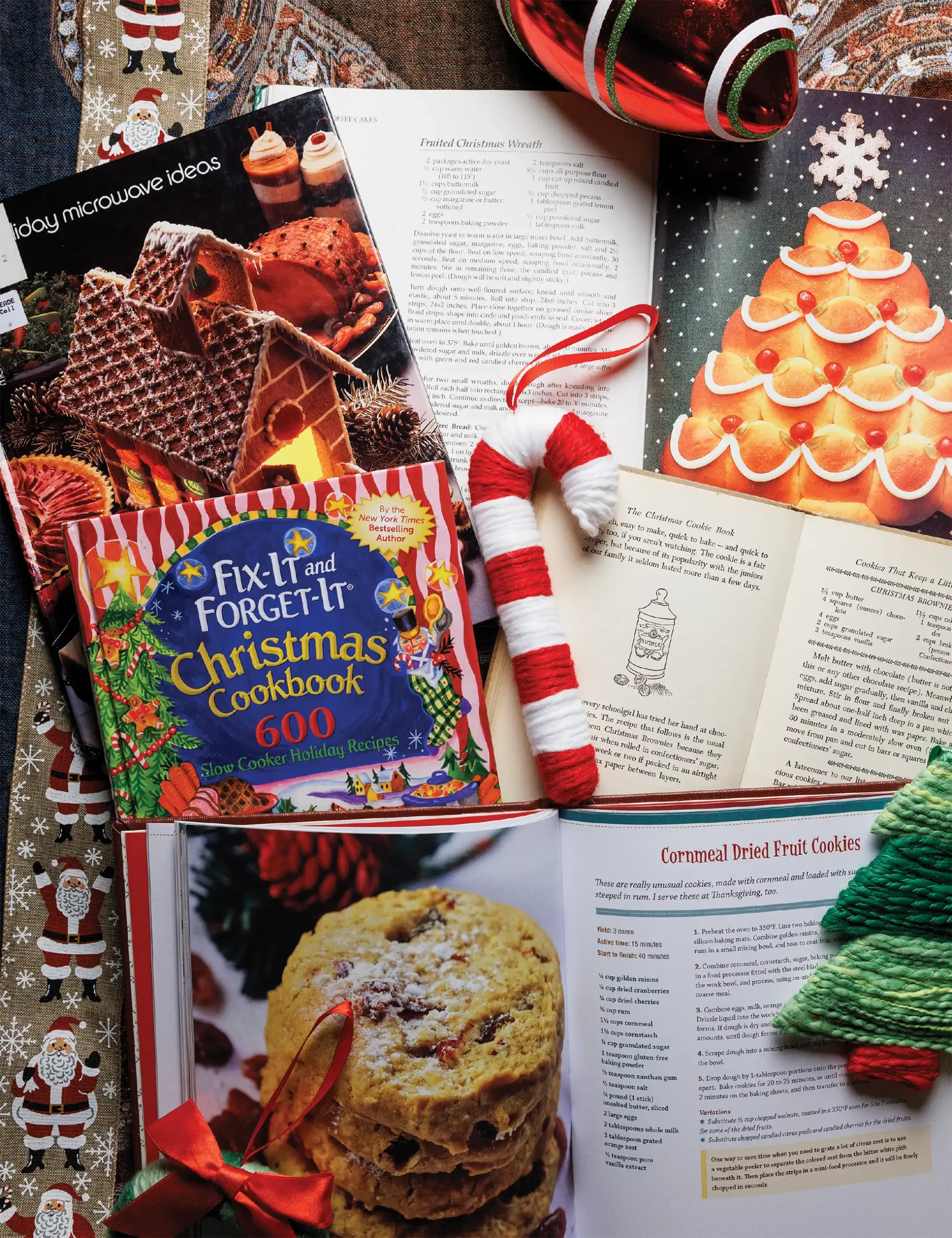 A collection of open holiday-themed cookbooks arranged on a festive table with various Christmas decorations, including a red-and-white striped candy cane and green Christmas tree ornament.