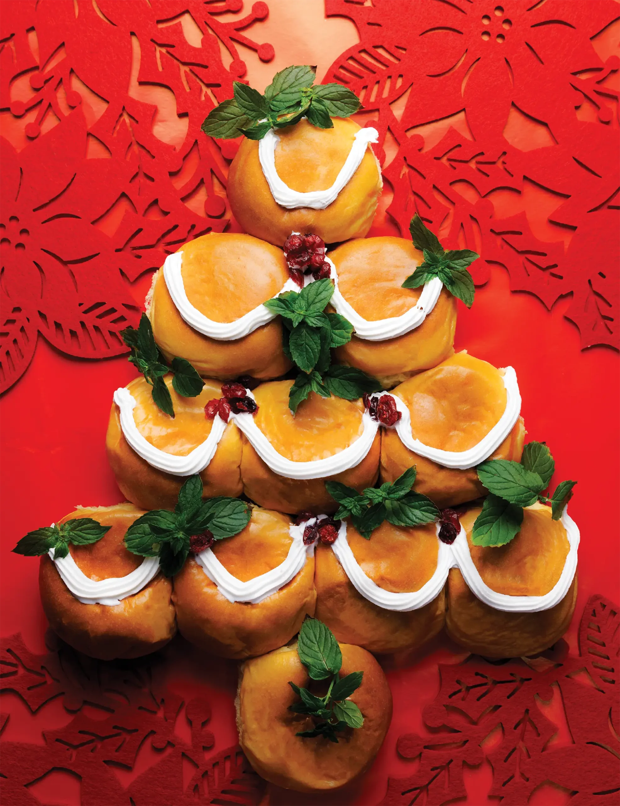 A holiday bread roll platter arranged in the shape of a Christmas tree, decorated with white frosting, mint leaves, and red cranberries on a red background with festive patterns.