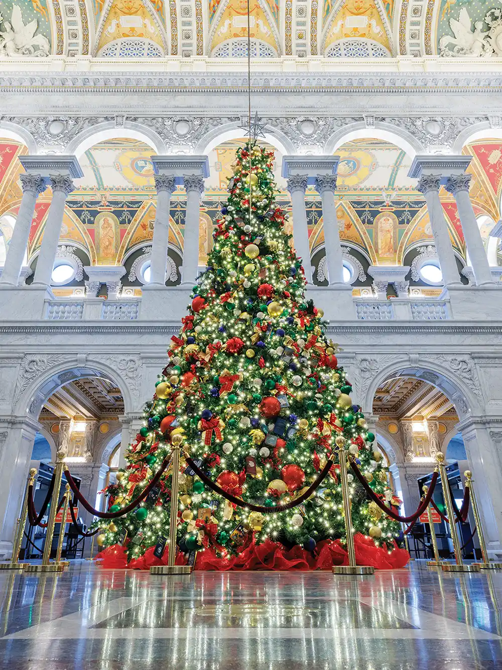 A large Christmas tree decorated with colorful ornaments and lights in an ornate building with marble columns and arches.