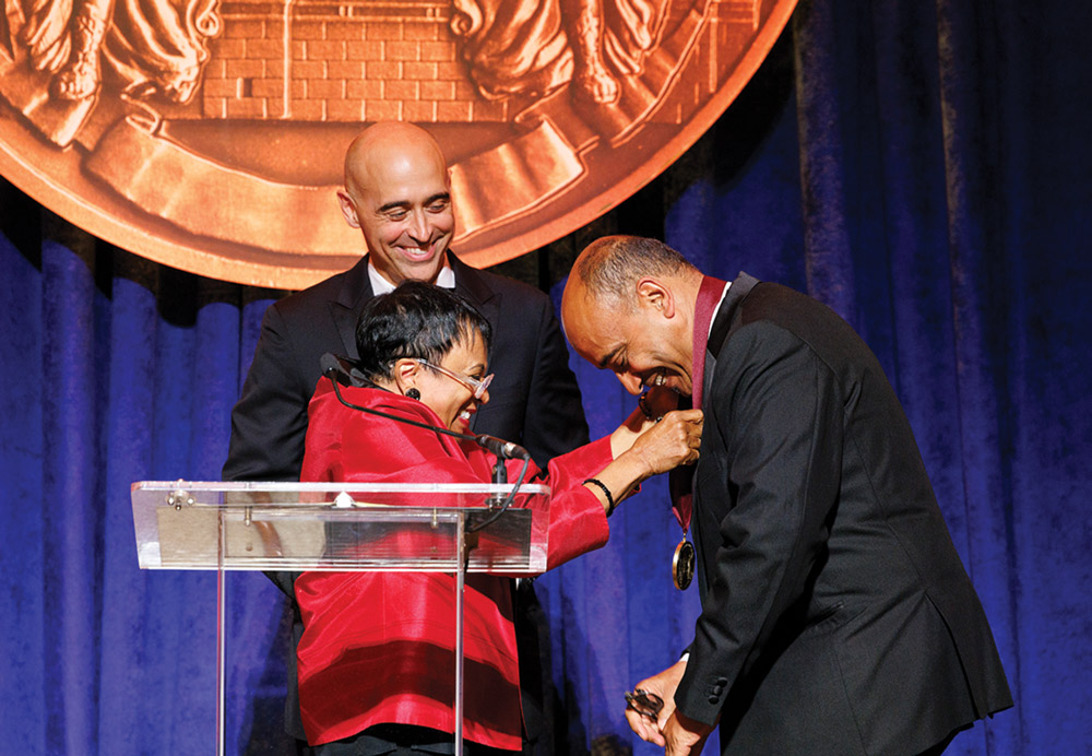 A woman awards a medal to a man on stage, while another man smiles in the background.