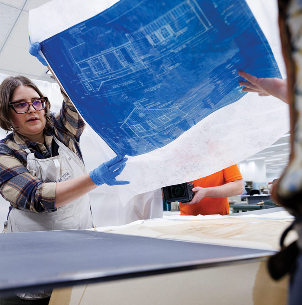 A woman holds up a translucent sheet with a blueprint underneath in an indoor workspace.