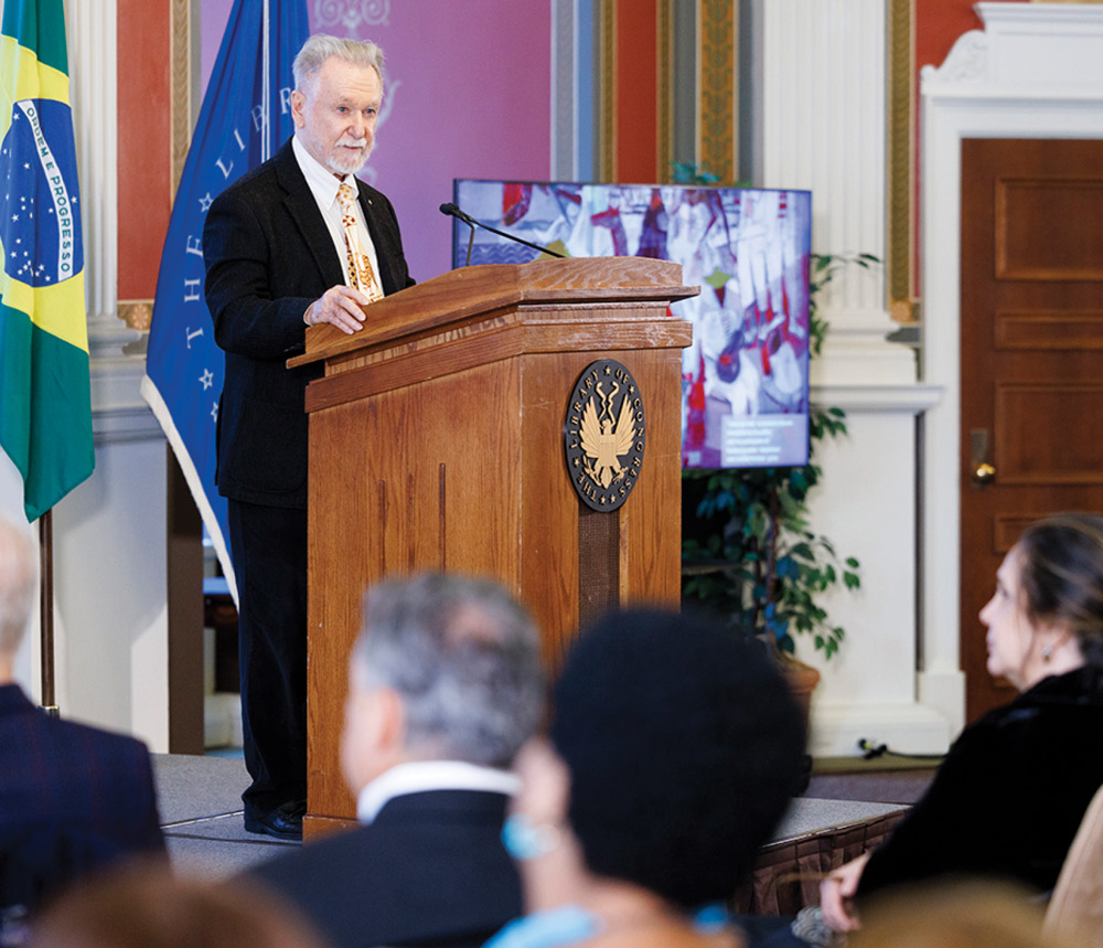 A man speaks at a podium with the Library of Congress emblem, Brazilian flag in background, addressing an audience.