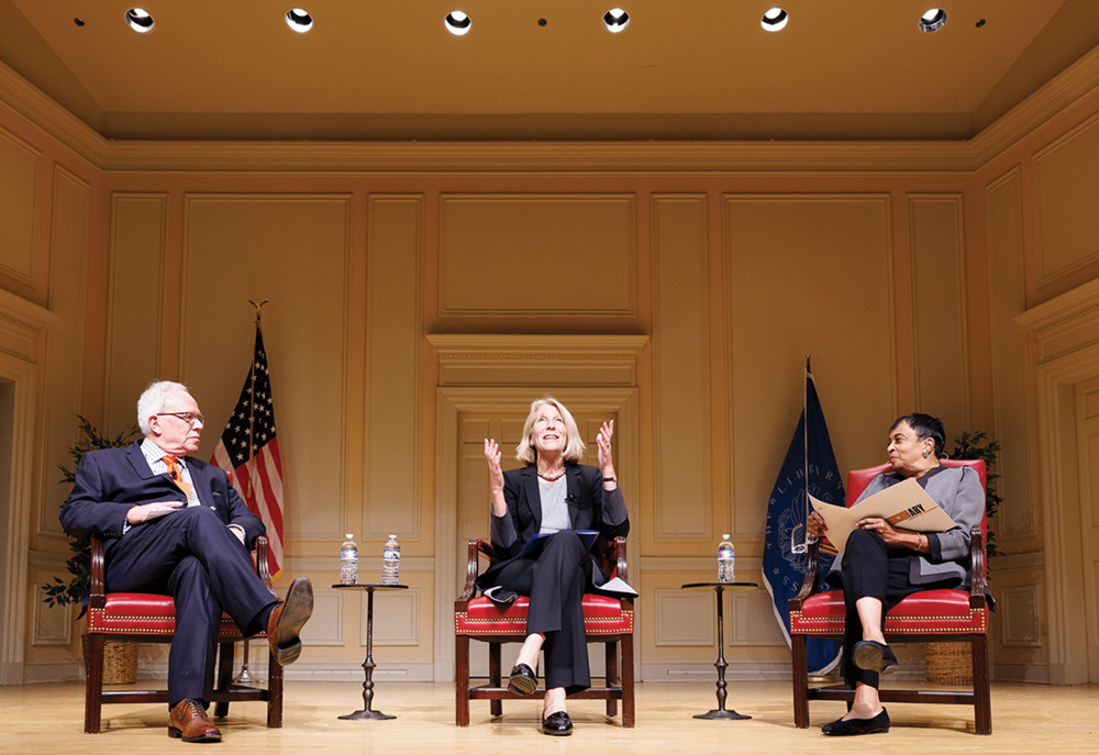 Three people seated in a panel discussion on stage with flags in the background.