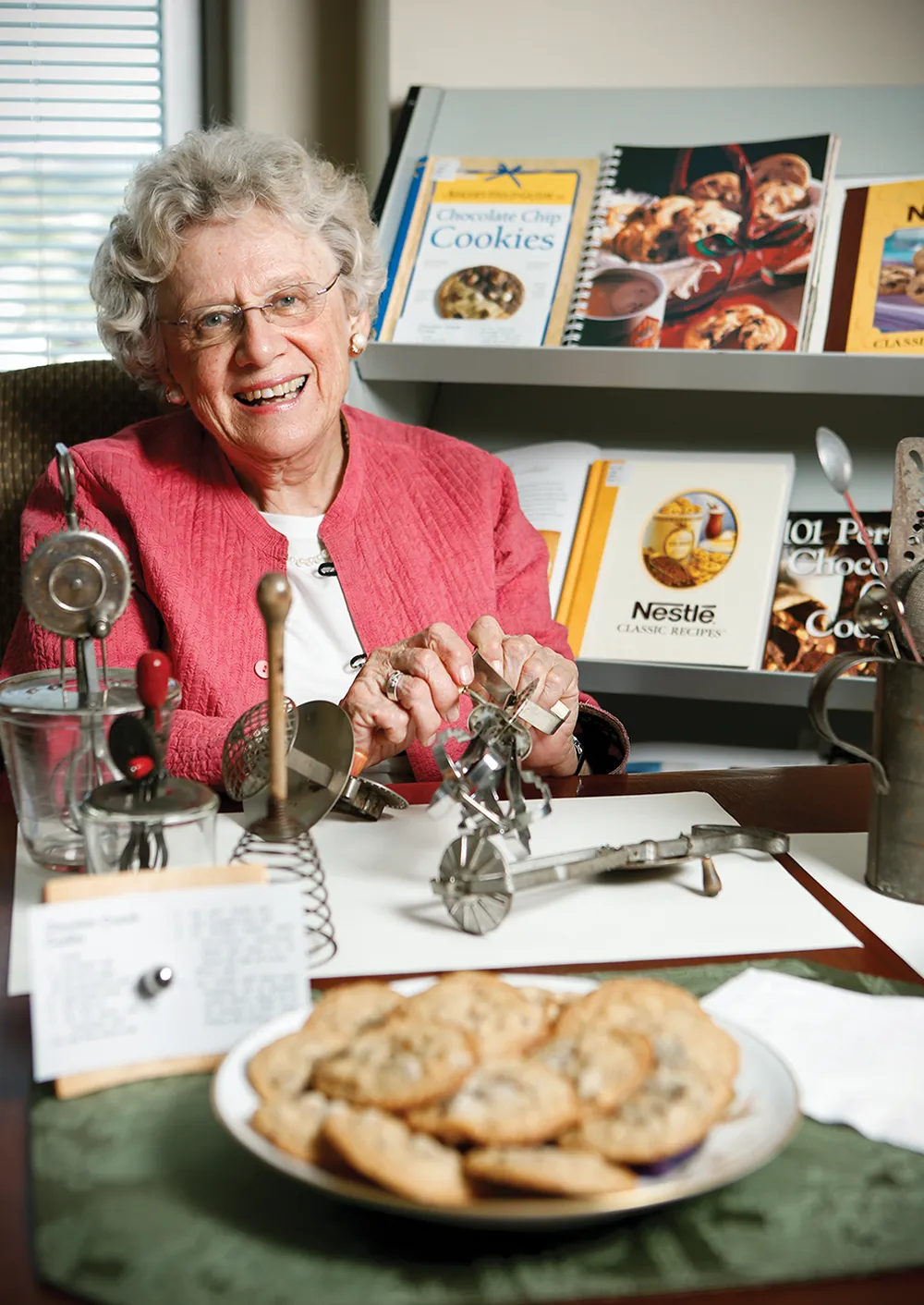 Connie Carter in pink jacket sitting at a table with vintage kitchen tools and cookies, with cookbooks in the background.