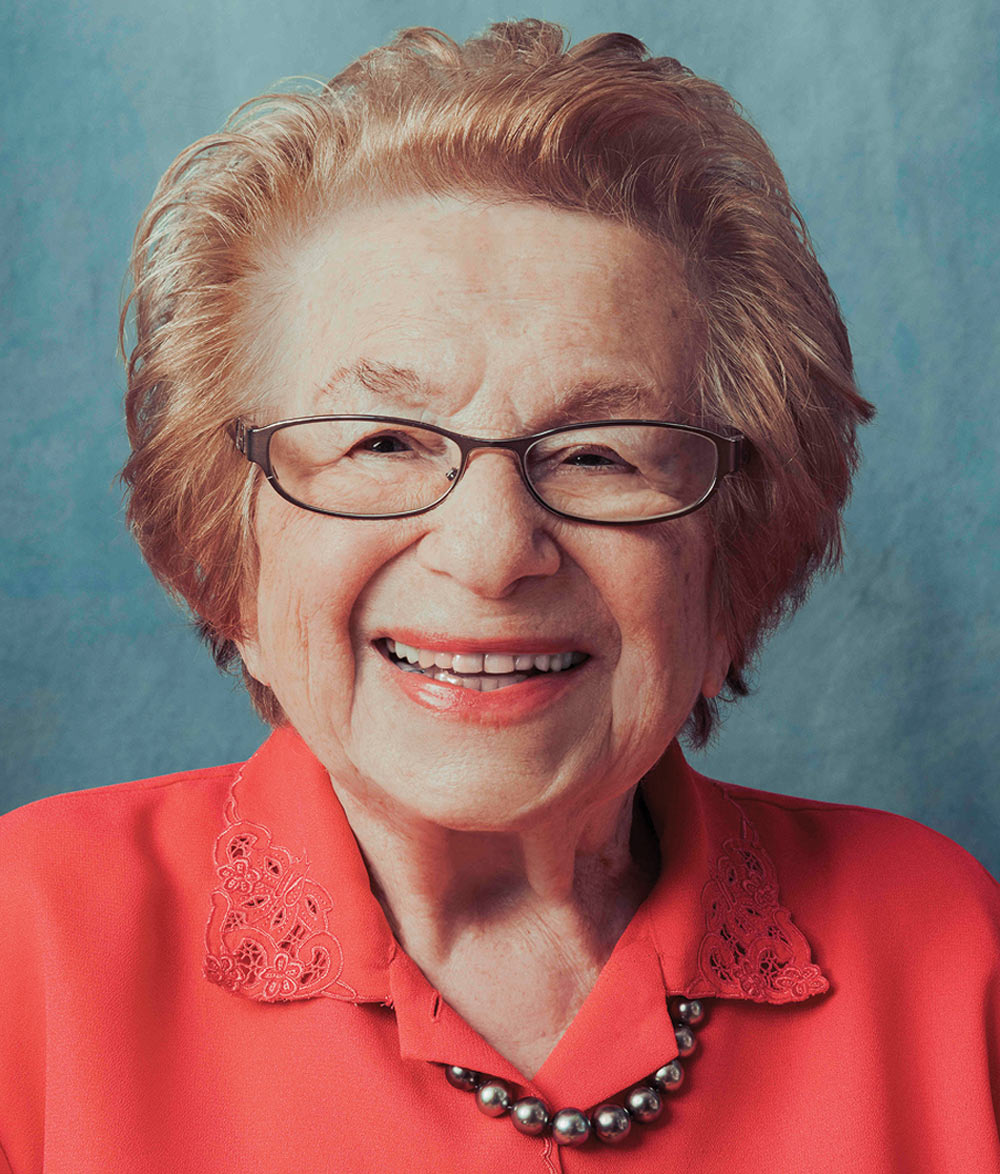 Ruth Westheimer with glasses smiling, wearing a coral blouse and pearl necklace against a soft blue background.