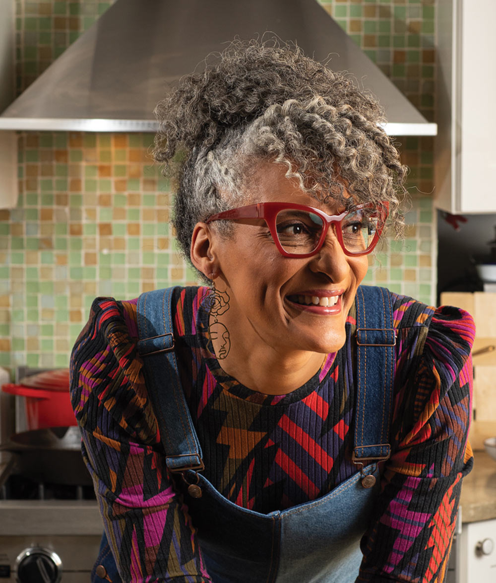 Carla Hall wearing red glasses and a patterned shirt in a kitchen setting.